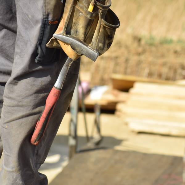 Beine einer Person in Arbeitskleidung, mit angehängtem Hammer, Blickrichtung auf einen Stapel mit Holzbrettern
