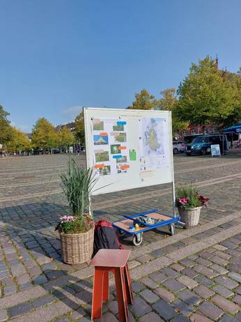 Whiteboard mit Infomaterialien, Blumenkübel daneben auf dem Großflecken in Neumünster