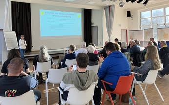 vorne sitzende Menschen mit Blick zur vortragenden Person, die hinten vor der Leinwand mit Präsentationsteht