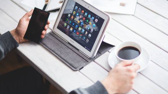 Symbolbild: Ein Mann sitzt vor einem Tablet, das auf dem Tisch steht. In der einen Hand hält er ein Smartphone, in der anderen Hand eine Tasse Kaffee.