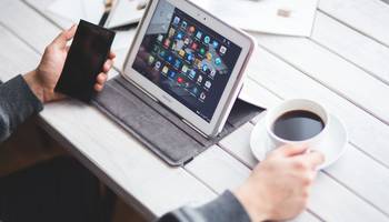 Smartphone- und Tablet-Sprechstunde Symbolbild: Ein Mann sitzt vor einem Tablet, das auf dem Tisch steht. In der einen Hand hält er ein Smartphone, in der anderen Hand eine Tasse Kaffee.