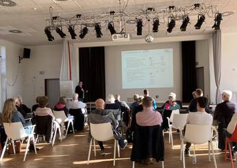 vorne sitzende Menschen mit Blick zur vortragenden Person, die hinten vor der Leinwand mit Präsentationsteht
